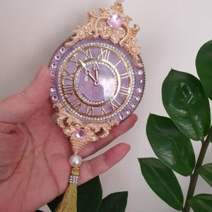 May include: A decorative clock with a lavender face and gold Roman numerals. The clock is adorned with rhinestones and gold filigree. A gold tassel and pearl hang from the bottom. Pink gemstones are set at the top.