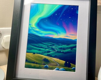 May include: A framed painting of a landscape with a winding road leading up a hill. The sky is a vibrant blue with a green, blue, and purple aurora borealis. The hills are green and the road is dirt.