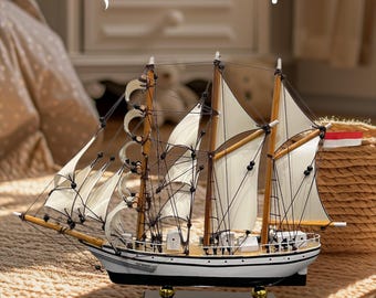 May include: A white and black wooden model ship with sails, a wooden base, and the text "The Warwick" on the side. The ship is on a beige knitted surface. The text "JUST IN!" is in the background.