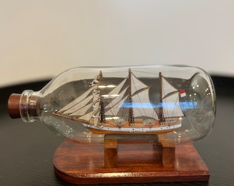 Op de afbeelding: Een miniatuur houten scheepsmodel in een heldere glazen fles, met een houten voet. Het schip heeft witte zeilen en de tekst "DE WARUS" is zichtbaar aan de zijkant.
