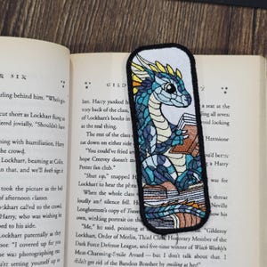 Puede incluir: Marcapáginas bordado con un dragón azul y turquesa leyendo libros. El dragón tiene detalles amarillos y un fondo blanco con borde negro. El marcapáginas está en un libro abierto.
