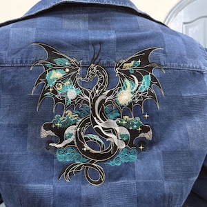 以下が含まれることがあります： 背中に刺繍されたドラゴンデザインが施された青いデニムジャケット。 ドラゴンは黒色で、銀色のアクセント、ターコイズとゴールドのハイライトがあり、雲と星を背景にしています。