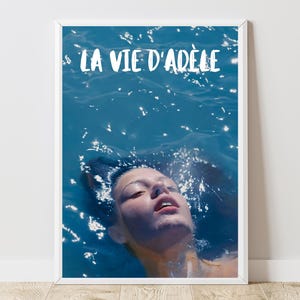 Puede incluir: Una impresión enmarcada con el texto "LA VIE D'ADÈLE" en blanco, que representa a una persona en agua azul. La imagen evoca una atmósfera tranquila, con el rostro de la persona parcialmente visible sobre la superficie del agua.
