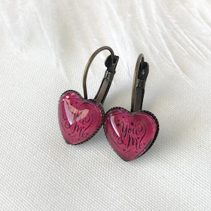 Puede incluir: Un par de pendientes en forma de corazón con un cabujón de vidrio rosa. Los cabujones presentan el texto "You & Me" en escritura negra.