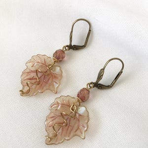 Pode incluir: Um par de brincos pendentes com um design de folha rosa e branca com detalhes em fio dourado. Os brincos têm um fecho de alavanca de bronze e uma conta de cristal rosa.