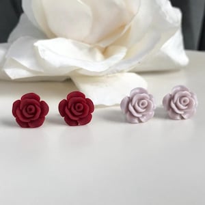 May include: Rose-shaped stud earrings in two colors. Two dark red earrings are on the left, and two light purple earrings are on the right. The earrings are in front of a large white flower.