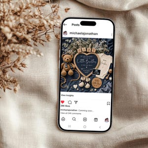 May include: A smartphone displays a baby announcement. The announcement includes a teddy bear, baby shoes, and a onesie. The text reads "Little Brother" and "October 2023". The background is a blue fabric with floral accents.