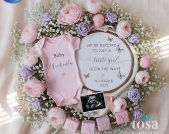 May include: A pink baby onesie with the name "Micheala" and a heart, surrounded by a floral wreath of pink and purple flowers. A circular sign reads "We're excited to say a little girl is on the way!" with the date November 2025.