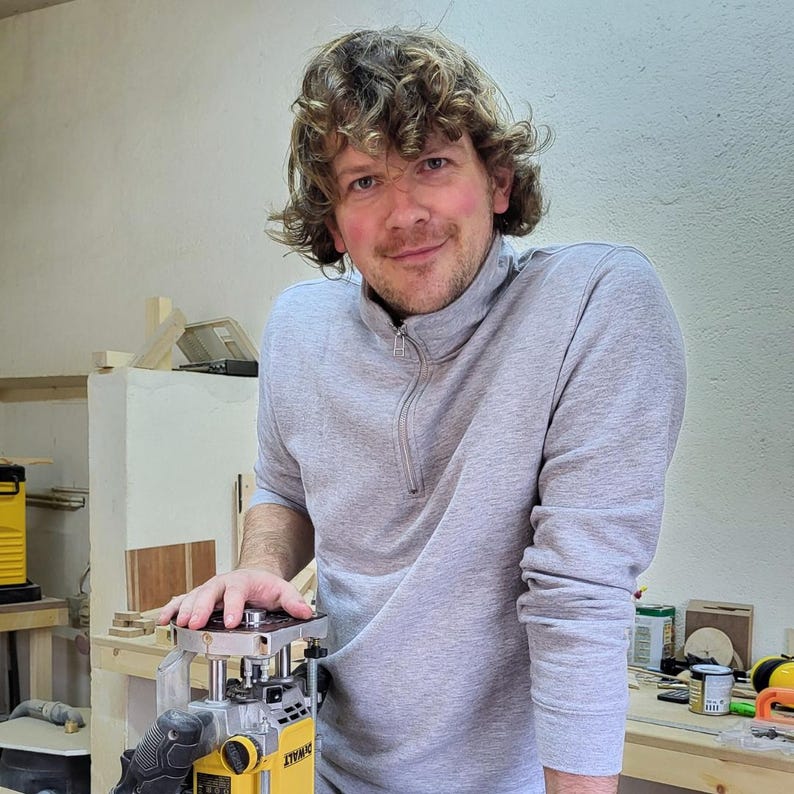 May include: A man wearing a gray sweater stands in a workshop, holding a yellow and black electric router. The router is on a wooden table with other tools and materials.