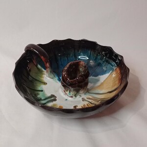 Peut inclure: Un bougeoir en céramique avec un glaçage brun, bleu, vert et blanc. Le bougeoir a une poignée et une base ronde.