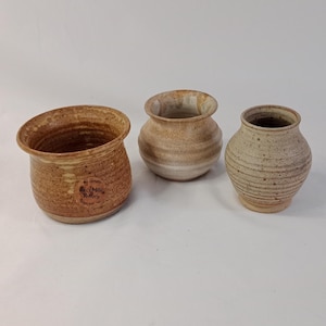 Puede incluir: Tres jarrones de cerámica hechos a mano de diferentes tamaños y formas. Los jarrones son principalmente de tonos tierra, con tonos marrones y beige. El jarrón de la izquierda tiene una abertura ancha y acampanada, mientras que los otros son más redondeados.