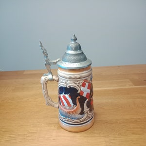 May include: A ceramic beer stein with a metal lid and a handle. The stein features a colorful design of a ship with sails and a flag. The stein is on a wooden surface.