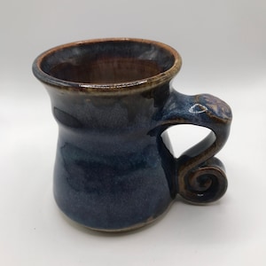 May include: A handmade ceramic mug with a blue glaze and a brown rim. The mug has a unique, curled handle.