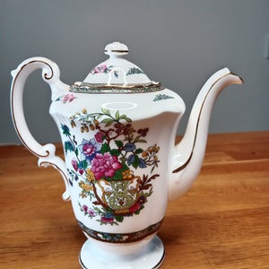 Peut inclure: Théière en porcelaine blanche avec un motif floral dans des tons de rose, jaune, vert et bleu. La théière est ornée d'une bordure dorée autour du couvercle et de la base.