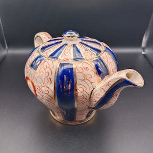 Può includere: Una teiera bianca con accenti blu e dorati. La teiera ha un corpo rotondo e un beccuccio curvo. Il design presenta motivi a spirale e strisce verticali.