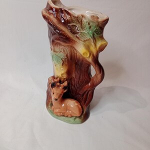May include: A ceramic vase shaped like a tree trunk with a deer figurine at the base. The vase is decorated with brown, green, and yellow paint.