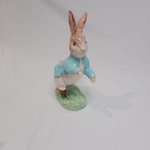 Puede incluir: Figura de cerámica de un conejo, con una chaqueta azul claro y zapatos rojos, de pie sobre una base verde. El conejo tiene orejas marrones y una cara blanca. Un artículo decorativo.