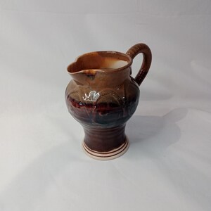 May include: A brown ceramic pitcher with a white rim and a brown handle. The pitcher has a textured surface and a rounded body.