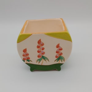 Könnte beinhalten: Ein weißer Keramiktopf mit grünen, gelben und orangefarbenen Blumendesigns. Der Topf hat drei kleine Füße und eine quadratische Öffnung oben.