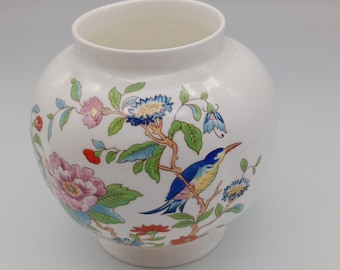 Puede incluir: Un jarrón de cerámica blanca con un diseño floral y un pájaro. El jarrón tiene un cuerpo redondo y una boca ancha. El diseño presenta flores rosas, azules, amarillas y verdes, así como un pájaro azul con detalles amarillos.