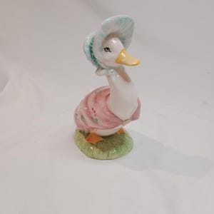 Puede incluir: Figura de pato de cerámica con un vestido rosa y un gorro azul. El pato tiene un pico amarillo y patas naranjas, de pie sobre una base de hierba verde. La figura está pintada con detalles delicados.