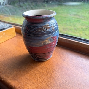 Pode incluir: Um pequeno vaso de cerâmica com corpo arredondado e borda alargada. O exterior apresenta uma superfície texturizada com linhas onduladas horizontais, com acabamento em tons de vermelho profundo, azul escuro e dourado metálico.