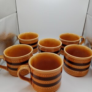 May include: Six brown ceramic mugs with a brown and black striped pattern. The mugs are arranged in a circle on a white surface.