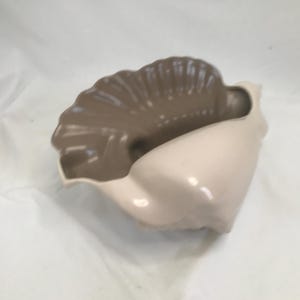 Puede incluir: Un cuenco decorativo de cerámica con forma de concha. El interior es de color marrón oscuro, mientras que el exterior es de color beige claro. El cuenco tiene un acabado brillante y está diseñado para parecerse a una concha.