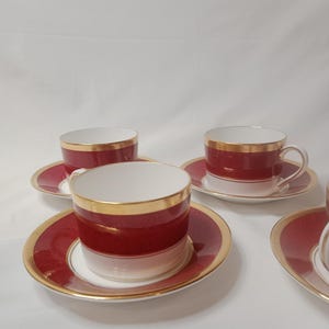 May include: Set of four red and gold teacups and saucers. The cups and saucers have a white rim and a gold band around the top.