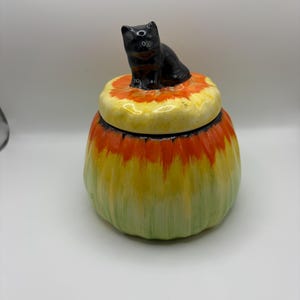 May include: A ceramic jar shaped like a pumpkin with a lid featuring a sculpted black cat. The jar has a gradient finish transitioning from a light green base to a bright yellow middle and an orange top, with a glossy, smooth surface texture.