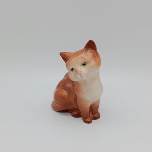 May include: A ceramic cat figurine, depicted in a seated pose. The cat features orange and cream-coloured fur and green eyes. The figurine is detailed with a glossy finish, presenting a classic cat pose.