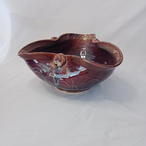 May include: A handcrafted, ceramic bowl with a rich, burgundy glaze. The bowl has an organic, undulating shape with a ruffled rim. Decorative elements include sculpted details and iridescent accents, creating a unique, artistic piece.