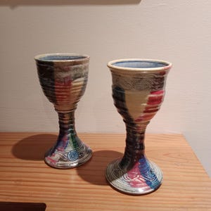 May include: Two handmade ceramic goblets with a colourful, abstract design. The goblets have a wide base, a slender stem, and a flared rim. The colours include blue, red, green, and brown. They are displayed on a wooden surface.