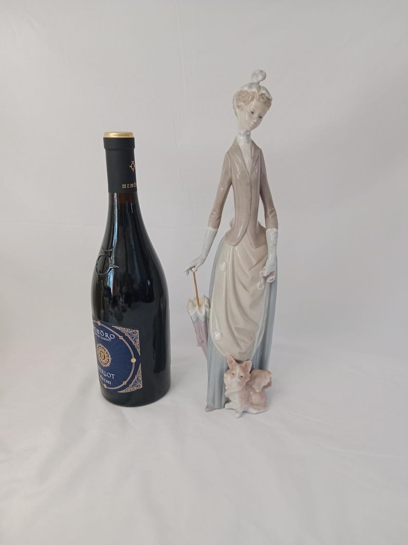 K&ouml;nnte beinhalten: Eine Porzellanfigur einer Frau in einem braunen Kleid, die einen Regenschirm und einen kleinen Hund h&auml;lt. Die Figur steht auf einer wei&szlig;en Oberfl&auml;che neben einer Weinflasche.