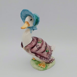 May include: A white porcelain figurine of a duck wearing a blue bonnet and a purple shawl with floral accents. The duck is standing on a green base with yellow feet.