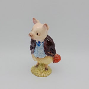 May include: A small ceramic figurine of a pig wearing a brown jacket, blue vest, and yellow pants. The pig is holding a red bag in its right hand and is standing on a green base.