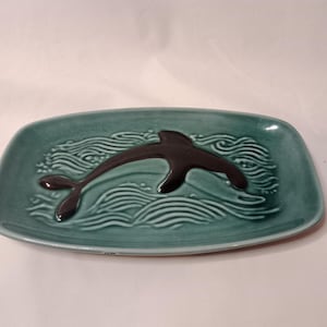 Könnte beinhalten: Eine tealfarbene Keramik-Servierplatte mit einem erhabenen schwarzen Delfin-Design. Der Delfin schwimmt über stilisierte Wellen.
