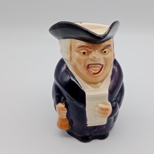 May include: A ceramic figurine of a man wearing a black hat and a dark purple coat. He is holding a scroll in his left hand and a bell in his right hand. The figurine is painted with realistic details, including a white face and a red mouth.