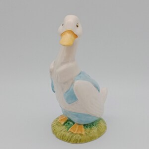 Puede incluir: Una figurita de pato de cerámica blanca con un chaleco azul y patas amarillas, de pie sobre una base verde.