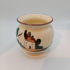 May include: A small, white ceramic pot with a brown rim and a painted design of a house with green trees. The pot is about 4 inches tall and 3 inches wide.