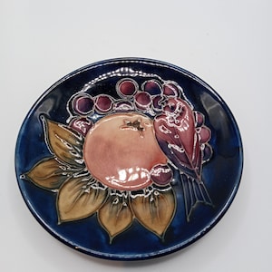 Puede incluir: Un plato de cerámica azul con un diseño en relieve de un pájaro posado sobre una fruta, rodeado de uvas. La fruta es un melocotón o una manzana de color rosa y naranja.