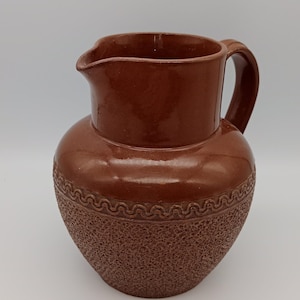 May include: A brown ceramic jug with a textured surface and a decorative band around the middle. The jug has a wide mouth and a handle.