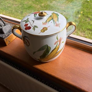 May include: A white porcelain lidded sugar bowl featuring gold-tone handles and trim. The exterior and lid are decorated with colourful illustrations of corn, cherries, and olives. The lid includes a small, round gold-tone knob handle.
