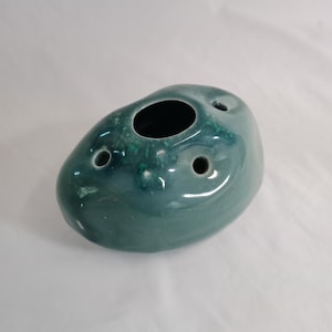 Puede incluir: Un jarrón de cerámica color turquesa con forma redondeada y orgánica. Tiene una gran abertura central y tres agujeros más pequeños. La superficie brillante refleja la luz, resaltando las variaciones de color.