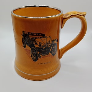 Puede incluir: Una taza de cerámica marrón con un borde plateado y una imagen en blanco y negro de un coche antiguo. El coche tiene una matrícula que dice "ARE 21" y el texto "LANCHESTER" está impreso debajo del coche.