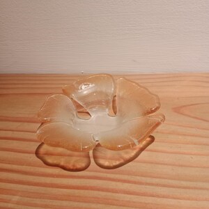 Könnte beinhalten: Eine blütenförmige Glasschale in hellem Orange. Die Schale hat eine klare Mitte und liegt auf einer Holzoberfläche. Die Blütenblätter sind durchscheinend und lassen Licht durch. Durchmesser ca. 15 cm.