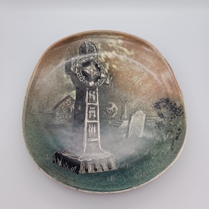 Könnte beinhalten: Ein Keramikteller mit handbemaltem Design eines keltischen Kreuzes auf einem Friedhof. Das Kreuz ist schwarz-weiß, der Hintergrund ist in gedeckten Grün- und Brauntönen gehalten.