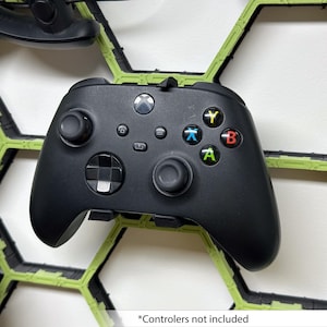 May include: A black Xbox controller with green, yellow, blue and red buttons is mounted on a green honeycomb wall mount. "Controllers not included" is written below the controller.