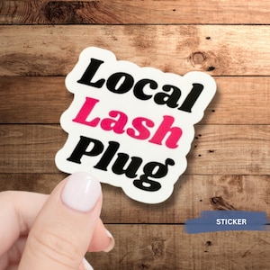 May include: A white sticker with the words "Local Lash Plug" in black and pink lettering. The sticker is held by a hand with a light pink nail polish. The background is a wooden surface.