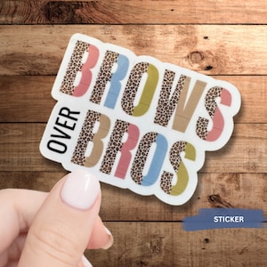 May include: A sticker with the text "Brows Over Bros" in colorful letters with a leopard print pattern. The sticker is on a wooden background.